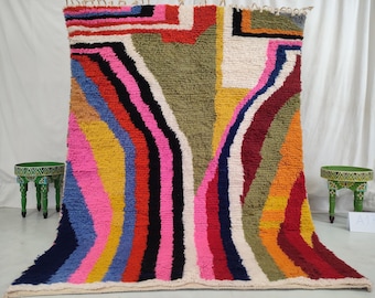 May include: A colorful, abstract rug with a geometric pattern in shades of pink, blue, yellow, red, black, and green. The rug has a white background and is made of wool.