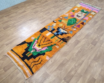 May include: A long, rectangular rug with a colorful geometric pattern. The rug is primarily orange with black, green, pink, and blue accents. The pattern includes triangles, diamonds, and other abstract shapes.
