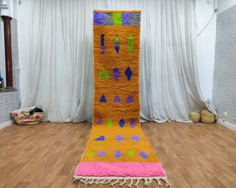 May include: A long, rectangular, orange rug with a pink border and a geometric pattern in shades of blue, green, and purple. The rug is made of wool and has a fringe on the ends.