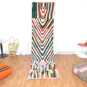 May include: A colorful Moroccan rug with a geometric pattern in shades of red, orange, green, and blue. The rug is hanging on a white wall in a room with a wooden floor.