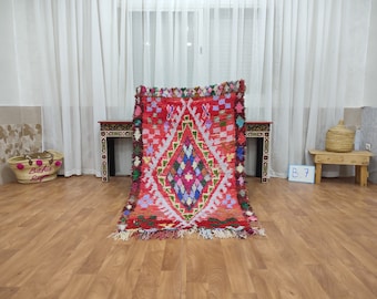May include: A red, white, blue, and pink Moroccan rug with a diamond pattern. The rug is laid out on a wooden floor in front of a white curtain.