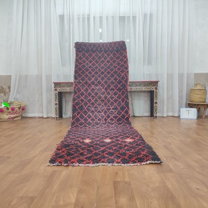 May include: A long, narrow, red and black Moroccan rug with a geometric pattern. The rug is laid out on a wooden floor in a room with white curtains.