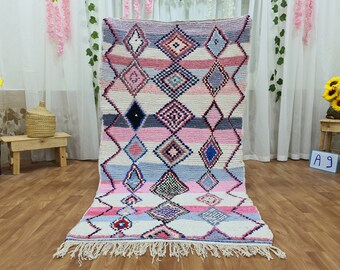 May include: A pink and blue Moroccan rug with a diamond pattern. The rug has a white background and is made of wool. The rug has a fringe on the bottom.
