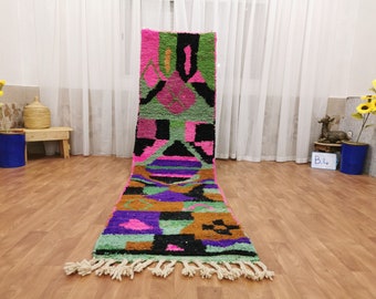 May include: A long, colorful rug with geometric patterns in shades of pink, green, purple, and black. The rug has a fringe on both ends.