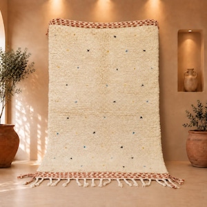 May include: A rectangular cream-colored wool rug featuring a shaggy texture, small scattered black, blue, and yellow tufted accents, and a decorative border at the top and bottom with a rust-colored checkered pattern and long fringe tassels.