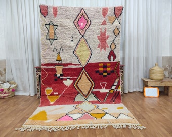 May include: A colorful Moroccan rug with geometric patterns in shades of pink, red, yellow, and white. The rug has a fringe border and is made of wool.