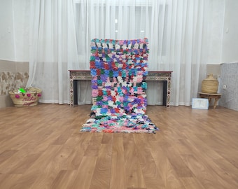 May include: A colorful Moroccan rug with a patchwork design in shades of pink, blue, green, and black. The rug is laid out on a wooden floor in a room with white curtains.