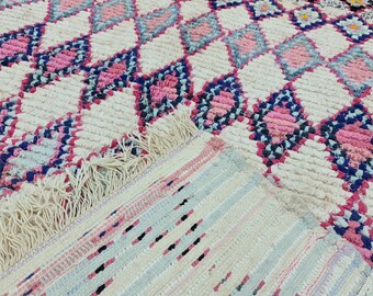 May include: A white, pink, and blue woven rug with a diamond pattern. The rug has a fringe border and is made of a soft, fluffy material.