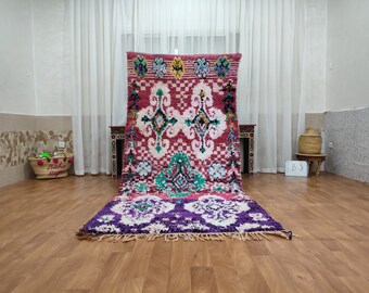 May include: A pink, white, and purple Moroccan rug with a geometric pattern. The rug has a fringe border and is laid out on a wooden floor.