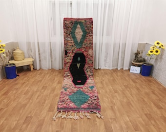May include: A colorful Moroccan rug with geometric patterns in shades of pink, blue, black, and white. The rug has fringe on the ends and is laid out on a wooden floor.