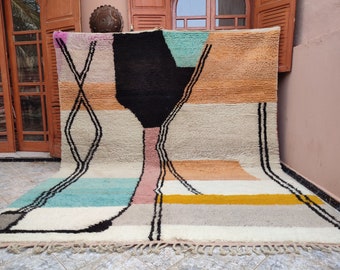 May include: A colorful abstract rug with geometric shapes in shades of pink, orange, yellow, blue, black, and white. The rug has a fringe border and is made of wool.
