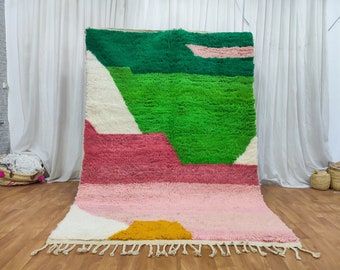 May include: A colorful abstract rug with geometric shapes in shades of green, pink, white, and yellow. The rug has a fringe border and is made of wool.