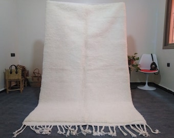 May include: A white shag rug with a fringe border. The rug is laid out on a grey floor in a room with white walls.
