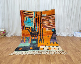 May include: A colorful Moroccan rug with geometric patterns in shades of orange, red, blue, green, and black. The rug has a fringe border and is made of wool.