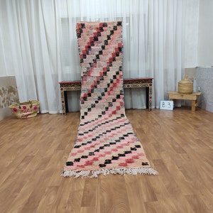 May include: A long, rectangular pink and black Moroccan rug with a geometric pattern. The rug is laid out on a wooden floor in a room with white curtains.