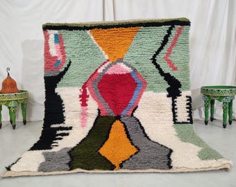 May include: A colorful Moroccan rug with geometric patterns in shades of green, red, orange, black, and white. The rug has a fluffy texture and is made of wool.
