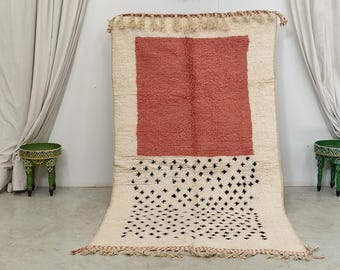 May include: A large, rectangular rug with a cream-colored background. It features a central, textured, coral-colored square and a lower section with a pattern of black cross-shaped designs. The rug has a fringe and tassels.