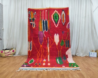 May include: A red Moroccan rug with a colourful abstract design featuring green, orange, pink, and blue accents. The rug has a fringe border.