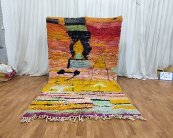 May include: A colorful Moroccan rug with a geometric pattern in shades of yellow, orange, pink, red, blue, and green. The rug has a fringe border and is laid out on a wooden floor.