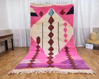 Puede incluir: Una alfombra marroquí rosa y blanca con un patrón geométrico. La alfombra tiene un borde de flecos y está hecha de lana.