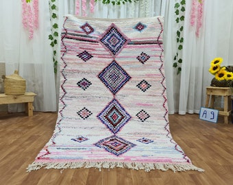 May include: A white Moroccan rug with pink, blue, and black diamond patterns. The rug has a fringe border.