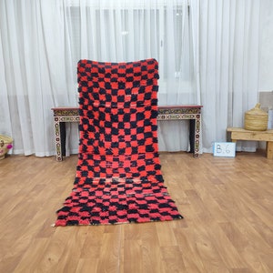 May include: A red and black checkered rug with a fringe border. The rug is laid out on a wooden floor in a room with white curtains.