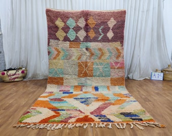 May include: A colorful Moroccan rug with geometric patterns in shades of orange, pink, blue, green, and white. The rug has a fringe border and is laid out on a wooden floor.