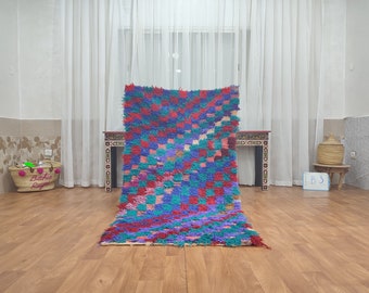 May include: A colorful Moroccan rug with a checkered pattern in shades of red, blue, green, and pink. The rug is laid out on a wooden floor in a room with white curtains.