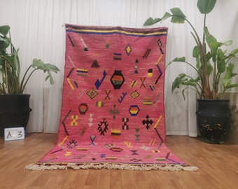May include: A pink Moroccan rug with a geometric pattern in shades of yellow, blue, black, and brown. The rug has a fringed border.