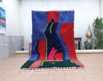Peut inclure: Un tapis abstrait coloré avec des formes géométriques dans des tons de bleu, rouge, vert, rose et noir. Le tapis a une bordure à franges et est fait de laine.
