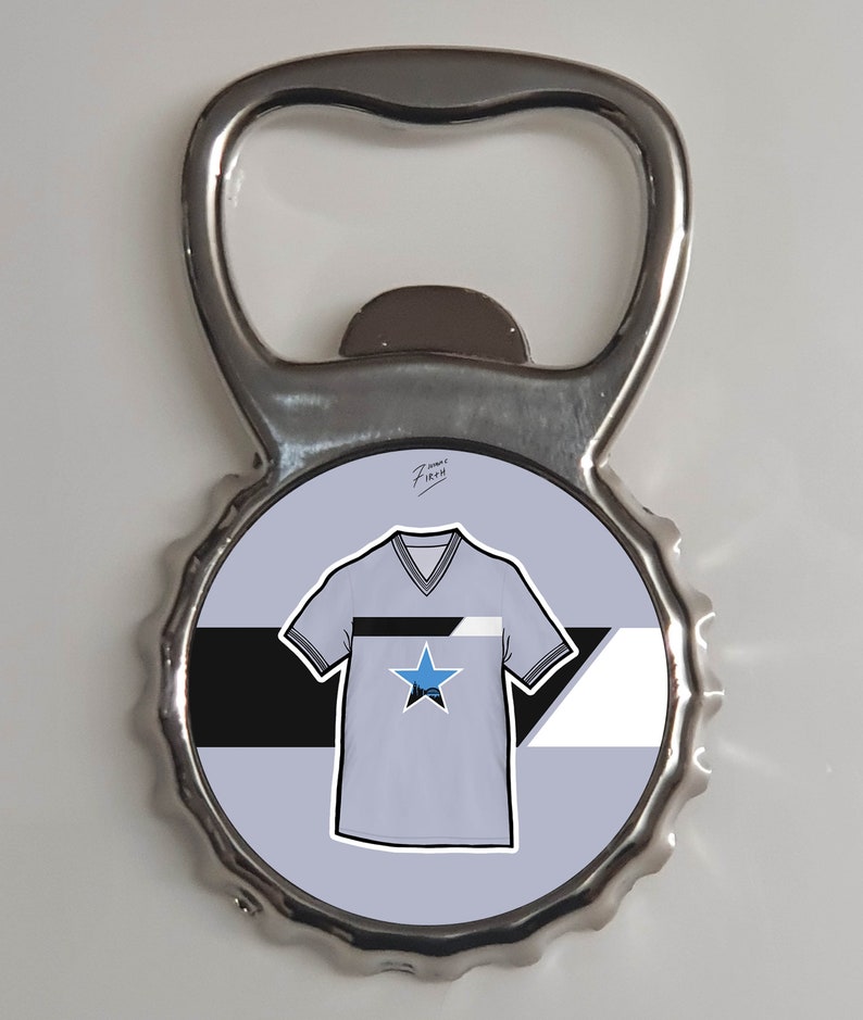 May include: A silver bottle opener with a round, blue, white, and black design featuring a stylized t-shirt with a star. The text "7 years FireH" is written in black on the design.