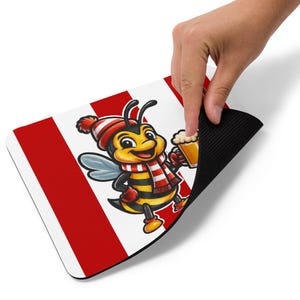May include: A rectangular mousepad featuring a cartoon bee wearing a red and white striped scarf and hat, holding a mug of beer. The background has red and white stripes. The mousepad is being held by a hand.