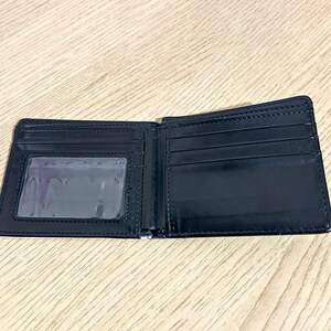 Puede incluir: Cartera negra plegable abierta, con una ventana transparente para identificaci&oacute;n y m&uacute;ltiples ranuras para tarjetas. La cartera est&aacute; hecha de un material liso y brillante. El interior est&aacute; vac&iacute;o.