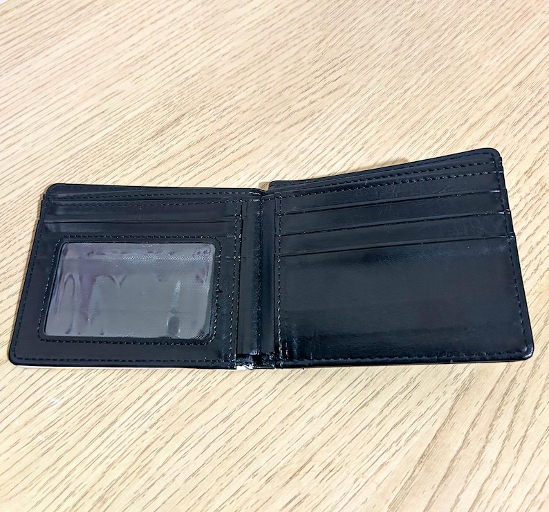 Puede incluir: Cartera negra plegable abierta, con una ventana transparente para identificaci&oacute;n y m&uacute;ltiples ranuras para tarjetas. La cartera est&aacute; hecha de un material liso y brillante. El interior est&aacute; vac&iacute;o.