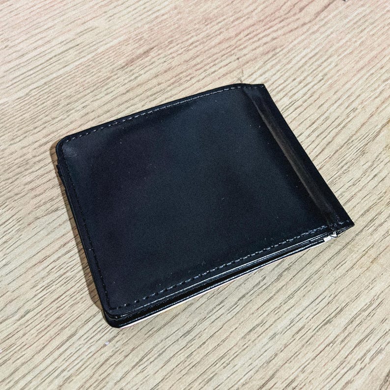 Puede incluir: Una cartera de cuero negro con un clip para billetes. La cartera rectangular tiene costuras blancas alrededor de los bordes. La cartera est&aacute; cerrada y parece vac&iacute;a.