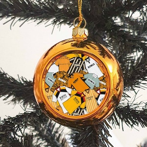 Puede incluir: Una bola de Navidad dorada brillante, con un surtido de camisetas deportivas amarillas, negras y blancas en su interior. La decoración cuelga de un hilo dorado y destaca sobre un árbol de Navidad verde oscuro.