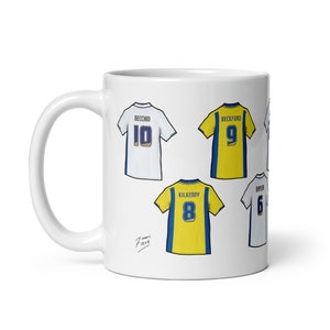 Può includere: Una tazza in ceramica bianca con un design di quattro maglie da calcio. Le maglie sono gialle e bianche con bordi blu. I numeri sulle maglie sono 6, 8, 9 e 10. I nomi sulle maglie sono Kilkenny, Beckford e Becchio.