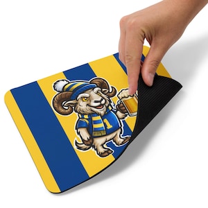 May include: A rectangular mousepad featuring a cartoon ram wearing a blue and yellow outfit, holding a beer. The background has blue and yellow vertical stripes. The mousepad is being held by a hand.