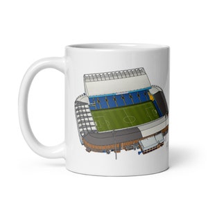 May include: White ceramic mug featuring a colourful illustration of a football stadium. The stadium has a green pitch, blue seats and a white roof.