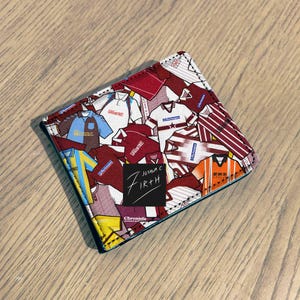 Peut inclure: Un portefeuille pliable avec un collage de maillots de sport de différentes couleurs, dont le bordeaux, le blanc, le bleu et l'orange. Le portefeuille porte le texte "Joshua C Firth" et "Chronicle" sur un carré noir.