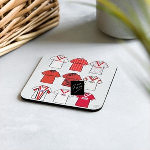 Peut inclure: Sous-verre carré avec des illustrations de maillots de football rouges et blancs. Le dessous de verre a une bordure noire et le texte "7 HOME FIRTH" dans un carré noir. Un panier en osier et un pot en céramique blanc sont en arrière-plan.