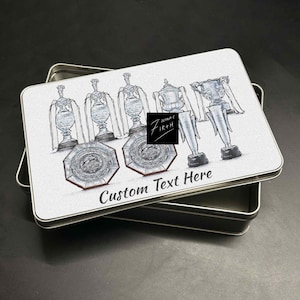 May include: A rectangular metal tin with a hinged lid, featuring a hand-drawn illustration of trophies and the text "Custom Text Here." The design is in shades of gray and silver, with a black logo in the center. The tin is open, revealing its interior.