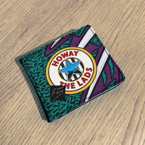 Może przedstawiać: Kwadratowy portfel z kolorowym wzorem. Zawiera napis "HOWAY THE LADS" w okrągłym emblemacie z niebieską gwiazdą. Projekt obejmuje fioletowe, zielone i białe geometryczne wzory. Widoczny jest mały czarny kwadrat z tekstem.