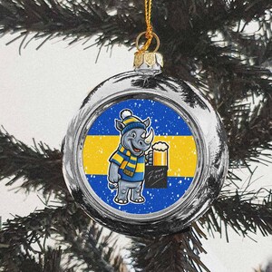 Puede incluir: Adorno navideño plateado con un rinoceronte de dibujos animados con gorro y bufanda azules y amarillos, sosteniendo una cerveza. El adorno tiene un fondo azul y amarillo y cuelga de un hilo dorado.