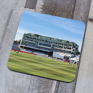 Puede incluir: Un estadio de cricket verde y blanco con un campo de hierba delante. Hay gente en el campo y en las gradas.