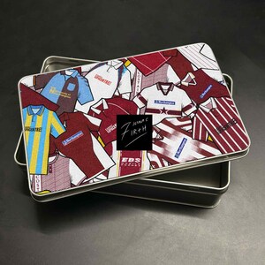 Peut inclure: Une boîte métallique rectangulaire avec un collage de maillots de football de différentes couleurs, dont le bordeaux, le bleu et le jaune. La boîte porte le texte "Joshua C Firth" et "EBS" sur un carré noir.