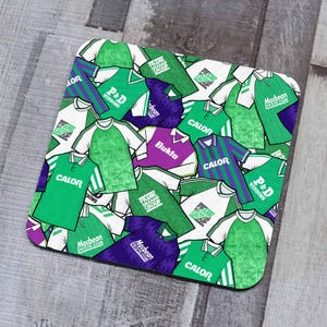 May include: A green and white patterned coaster featuring a collage of vintage football jerseys with various sponsors' logos, including "Calor", "P&D Windows", "Frank Graham Group", and "Macbean Protection Clothing".