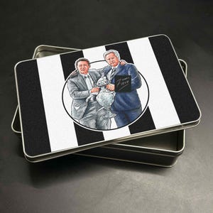 May include: A rectangular tin with a black and white striped design. The lid features a circular illustration of two men holding a trophy. The tin is open, revealing its interior.