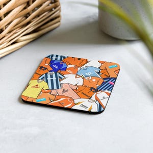 May include: Square coaster featuring a collage of illustrated soccer jerseys in various colors, including orange, blue, yellow, and white. The coaster has a black border and is placed on a white surface. The design includes text and logos.
