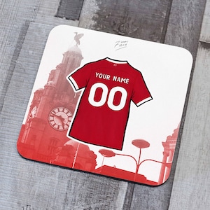 Puede incluir: Camiseta de fútbol roja y blanca con el texto "YOUR NAME" y el número "00" en la espalda. La camiseta está superpuesta sobre una foto de un edificio de ladrillo rojo con una torre de reloj.